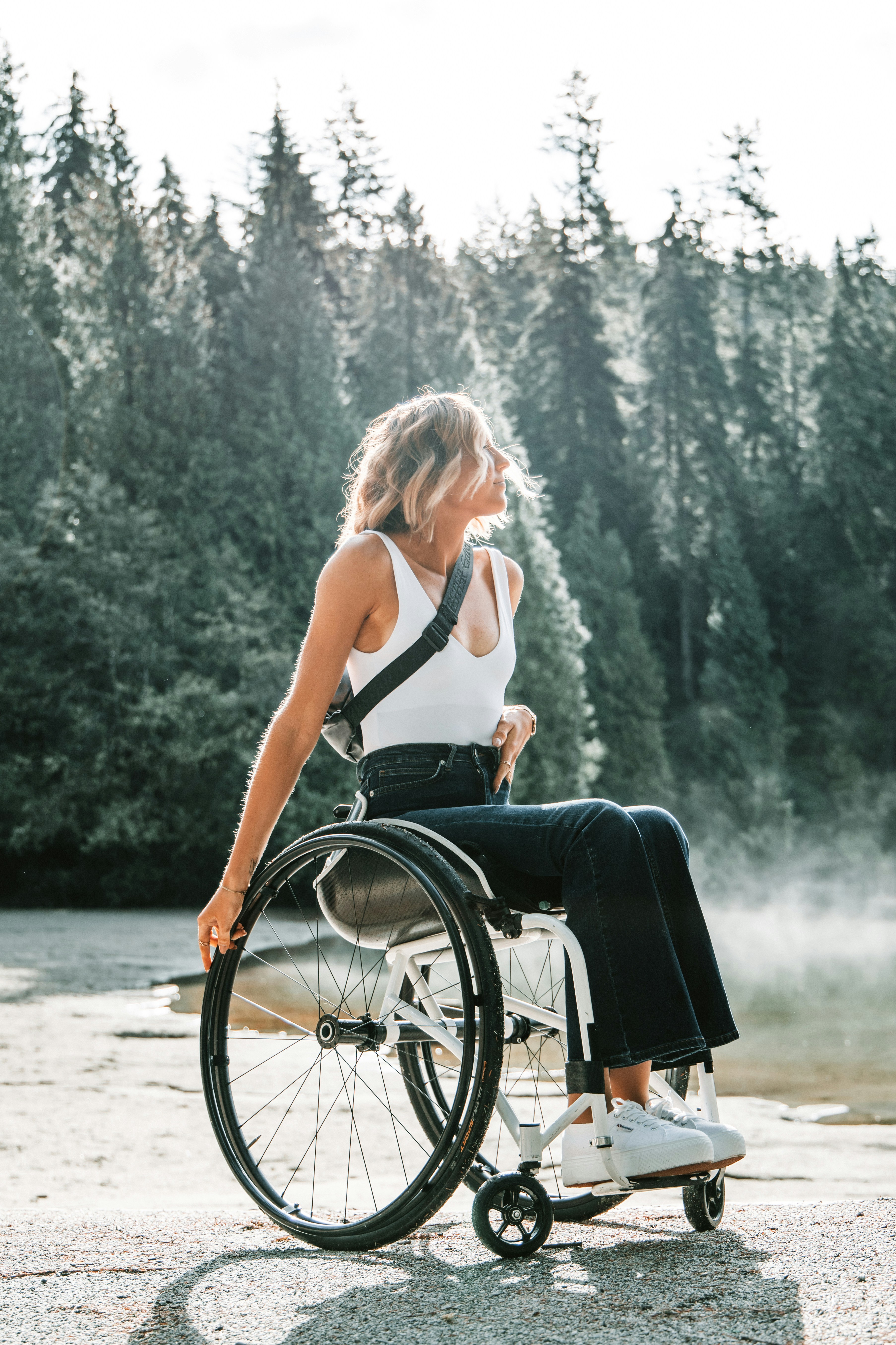 Jeune femme blonde en fauteuil roulant, vêtue d'un débardeur blanc et d'un jean, regardant vers la droite, face à un lac brumeux et une forêt de conifères.
