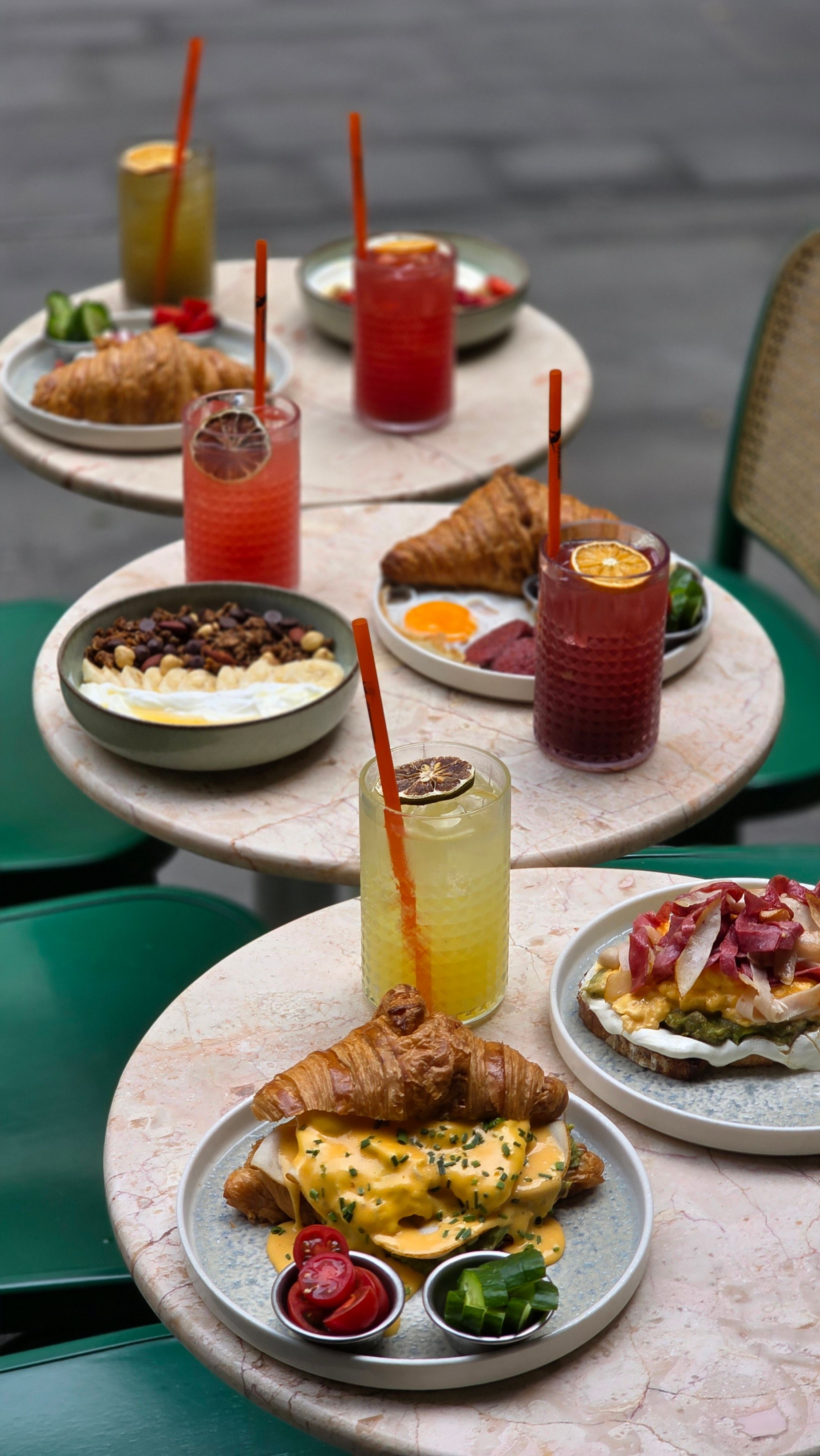 Tables de café avec divers petits déjeuners et boissons colorées : croissants, bols de granola, toasts, jus.